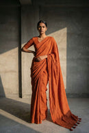 Woman in an orange saree standing against a plain wall.