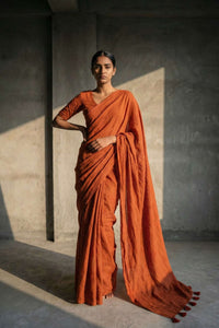 Woman in an orange saree standing against a plain wall.
