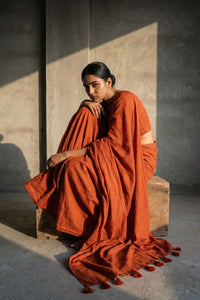 Woman in a rust-colored saree sitting on a wooden block against a concrete wall.