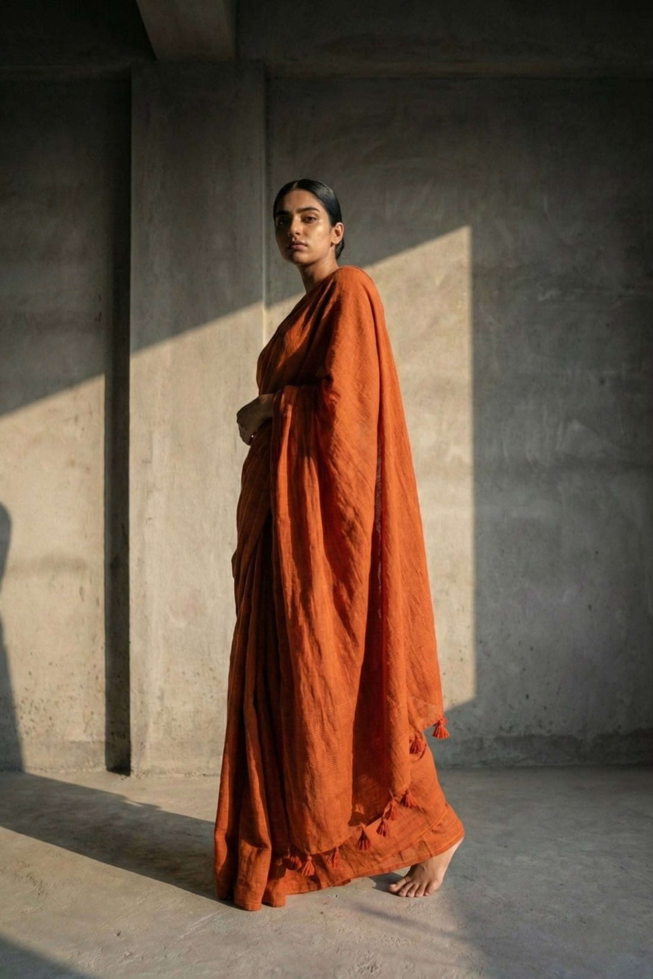 Woman in an orange saree standing against a textured wall with sunlight casting shadows.