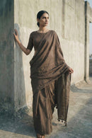 Woman in a brown saree standing against a concrete wall.