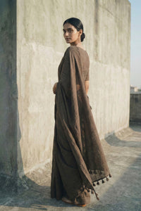 Woman in a brown saree standing against a textured wall.