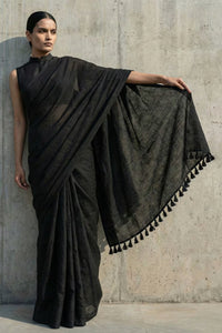 Woman wearing a black saree with tassels against a concrete wall.
