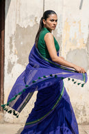 "Dynamic shot of a woman spinning, causing the indigo blue mul cotton saree and its tasseled pallu to flare out in motion. She is styled in a sleek high ponytail and a sleeveless green blouse. The background features a distressed, peeling beige wall that contrasts with the vibrant fabric."