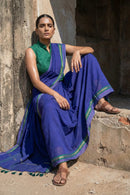"Full shot of a model sitting casually on rustic concrete steps against a weathered wall. She wears a deep indigo blue handloom saree with a green and gold border, paired with a high-neck sleeveless green blouse. She is wearing traditional brown leather sandals and maintains a serious, commanding expression."