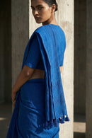 Woman wearing a blue saree standing against a concrete wall.