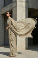 Woman in a beige saree standing against a concrete building.