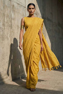 Woman wearing a mustard yellow saree against a concrete wall.