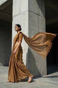 Woman in a brown mul cotton saree standing against a concrete wall.