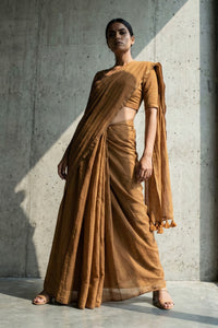 Woman wearing a brown mul cotton saree standing against a concrete wall.