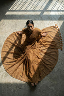 Woman in a flowing brown mul cotton saree on a concrete floor with sunlight casting shadows.