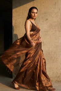 Woman in a flowing brown saree against a textured wall.
