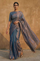 Woman wearing a metallic blue tissue saree against a beige wall.
