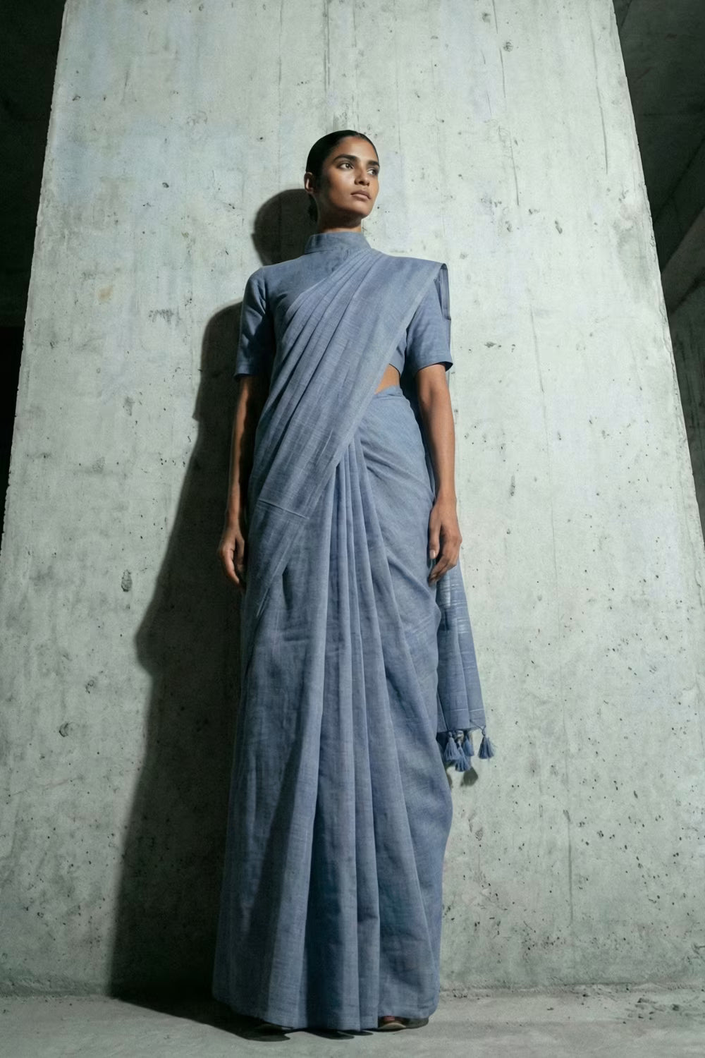 Woman wearing a blue saree standing against a concrete wall.