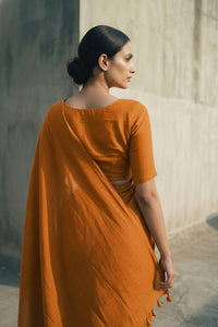Woman in an orange saree standing against a textured wall.