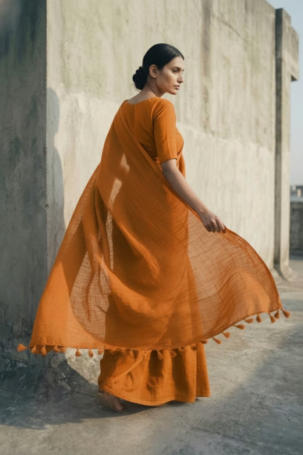Woman in an orange saree standing against a concrete wall.