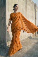 Woman in an orange saree standing against a concrete wall.