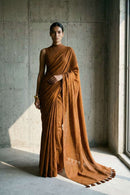 "Full-length editorial shot of a tall woman standing in a minimalist industrial space with raw concrete walls. She wears a cinnamon-brown handloom saree draped elegantly, with the long pallu trailing on the floor to reveal distinct tassel details on the hem."