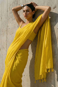 Woman in a yellow ml cotton saree leaning against a textured wall.