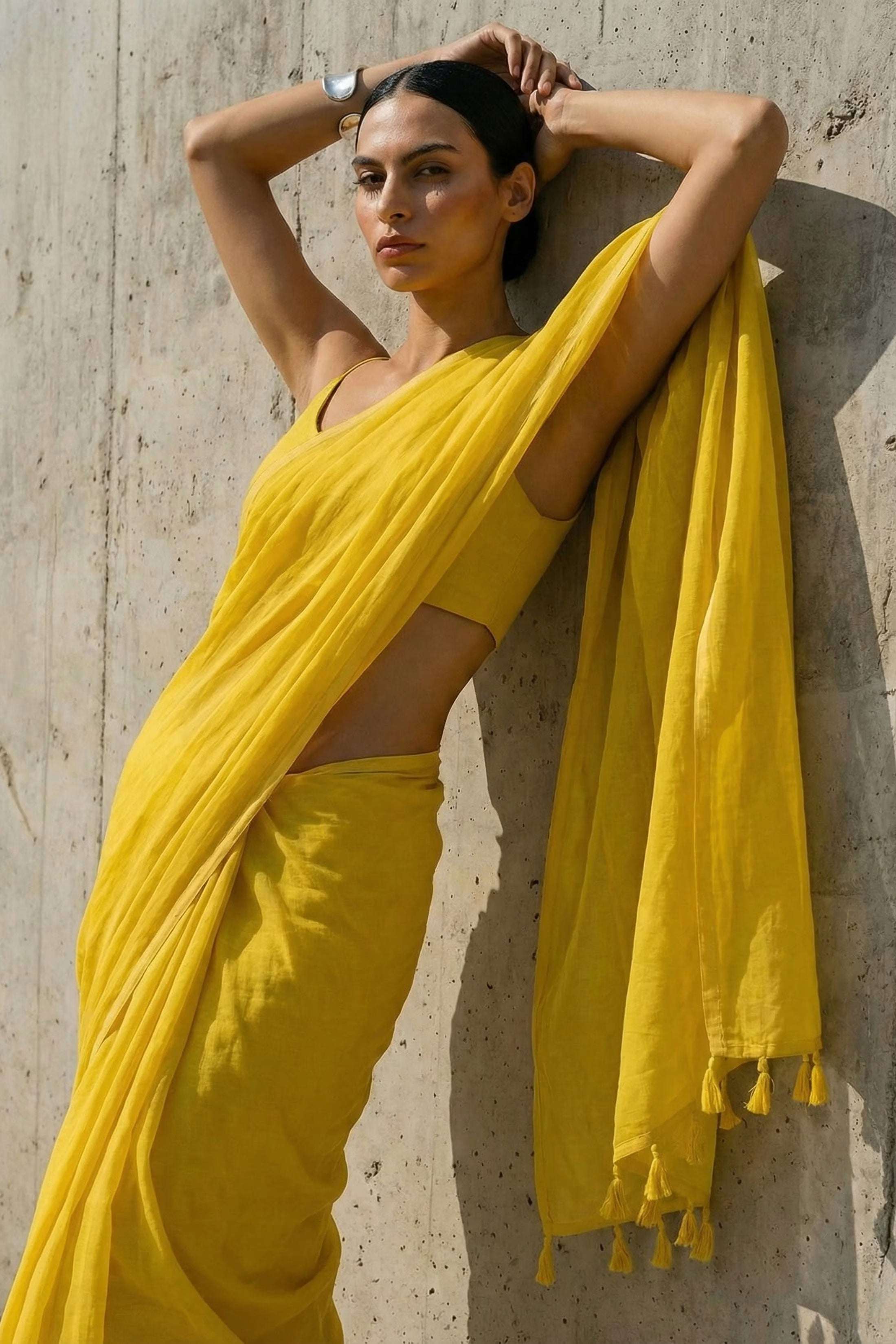 Woman in a yellow ml cotton saree leaning against a textured wall.