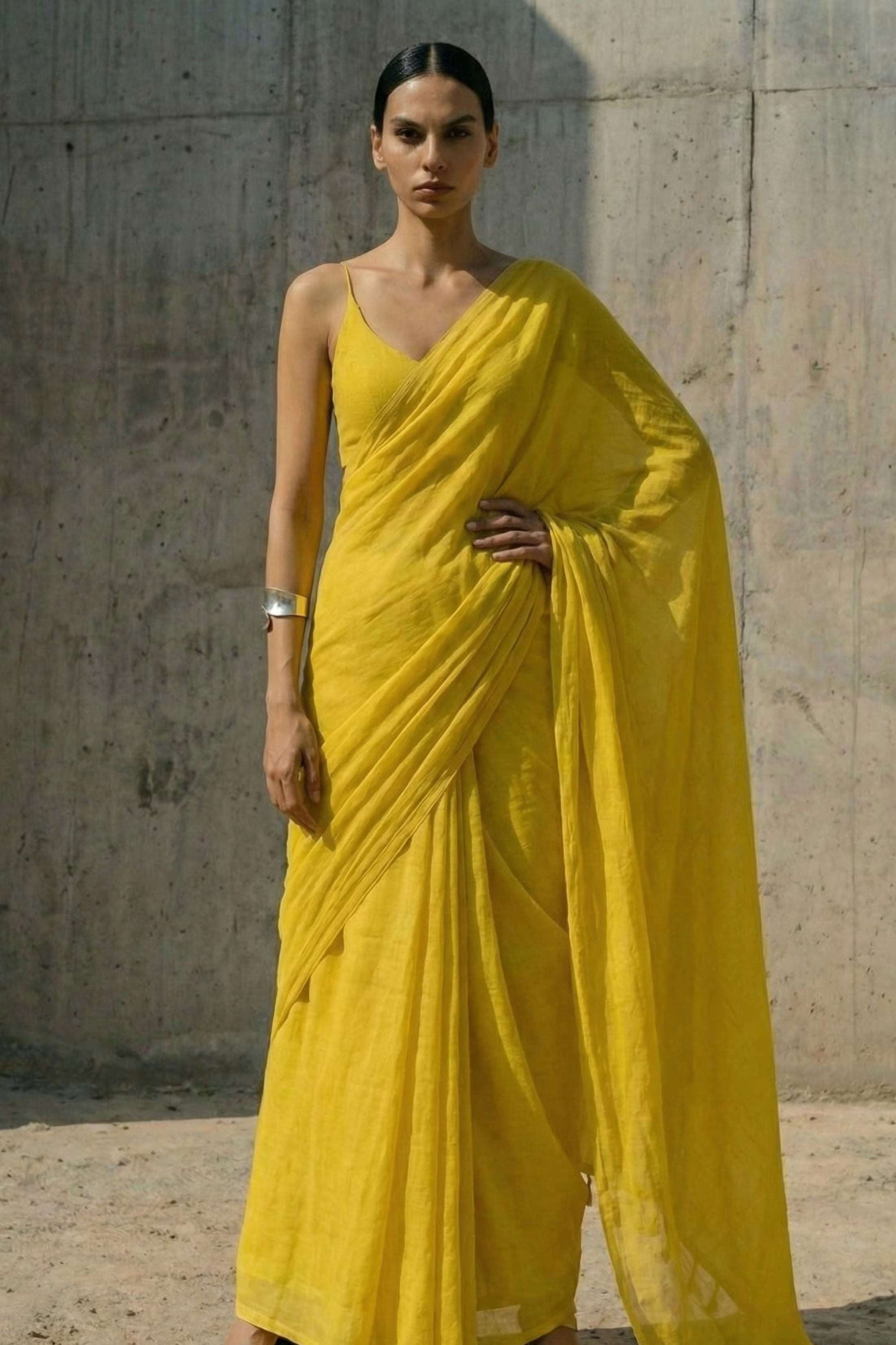 Woman wearing a yellow mul cotton  saree against a textured wall.