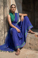 "Full shot of a model sitting casually on rustic concrete steps against a weathered wall. She wears a deep indigo blue handloom saree with a green and gold border, paired with a high-neck sleeveless green blouse. She is wearing traditional brown leather sandals and maintains a serious, commanding expression."