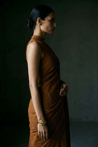 Side profile close-up of a South Asian model with sharp features and a sleek low bun. She is wearing a monochromatic rust-brown saree with a high-neck sleeveless blouse. A chunky gold cuff bracelet accents her wrist against a moody, dark grey textured background.