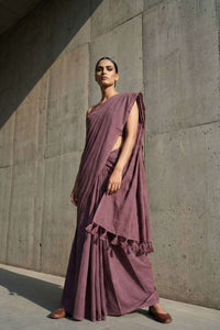 Woman wearing a purple saree standing against a concrete wall.