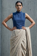 Woman wearing a blue top and beige saree against a concrete wall.