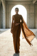 "Cinematic full-body shot of a model walking forward through a large concrete archway. She is backlit by bright natural light, creating a silhouette effect. Her rust-colored saree billows in the wind, dynamically displaying the flowing fabric and tassel accents."