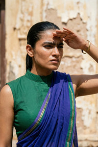 Woman in a green top and blue mul cotton saree with gold border against a stone wall background