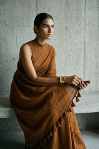 Medium shot of a model seated on a concrete ledge, wearing a monochromatic brown saree. She holds the edge of the fabric with both hands to display the decorative tassels on the pallu. The lighting is soft and natural, highlighting a gold statement cuff on her wrist.