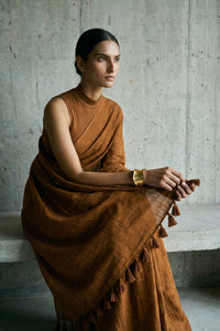 Medium shot of a model seated on a concrete ledge, wearing a monochromatic brown saree. She holds the edge of the fabric with both hands to display the decorative tassels on the pallu. The lighting is soft and natural, highlighting a gold statement cuff on her wrist.