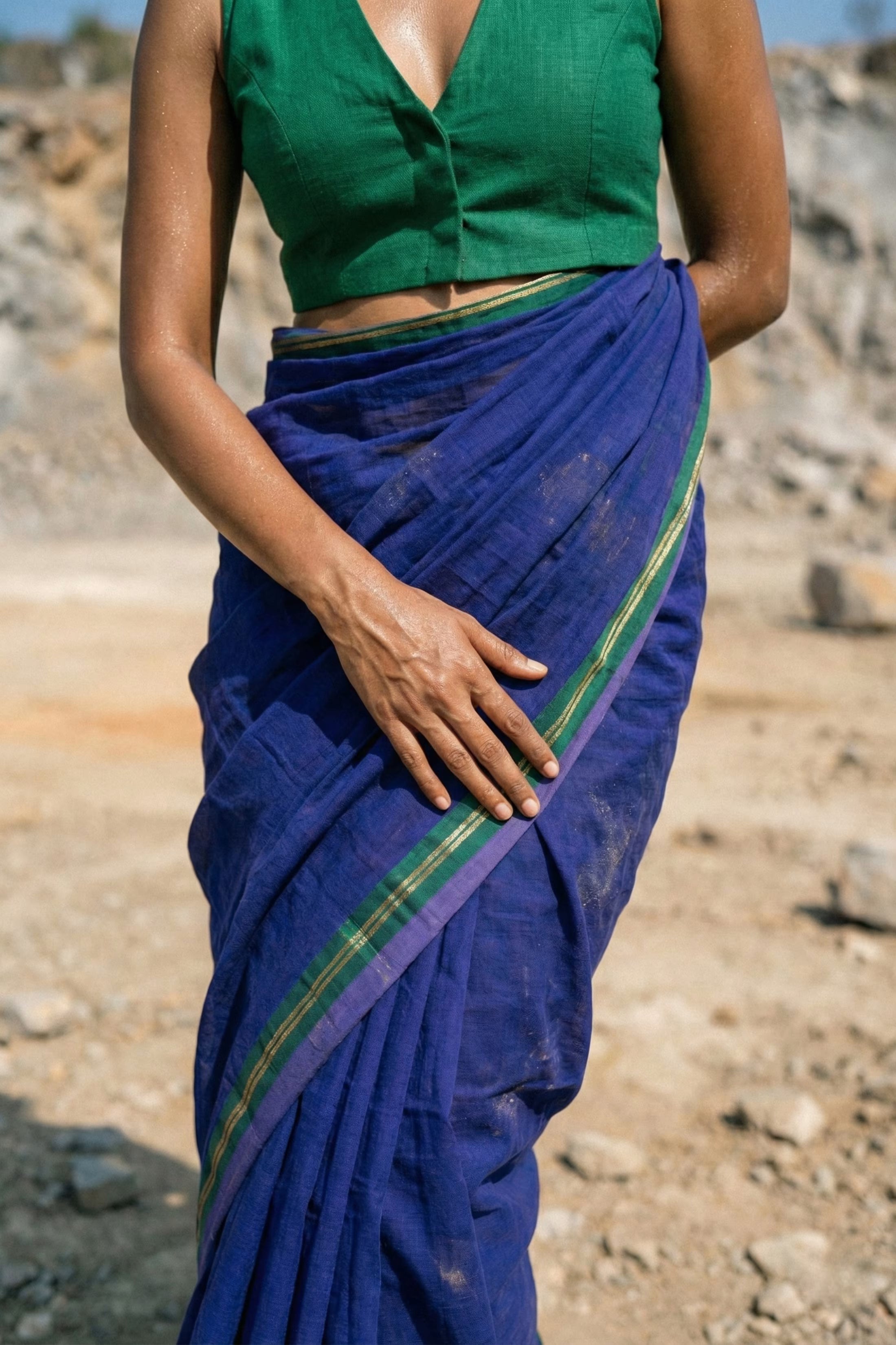 "Close-up crop of a model's torso highlighting the texture of a blue mul cotton saree with a green border. She wears a deep V-neck green blouse. The image captures a raw aesthetic with visible dust or sand on her skin and the fabric, emphasizing an outdoor, rugged setting."